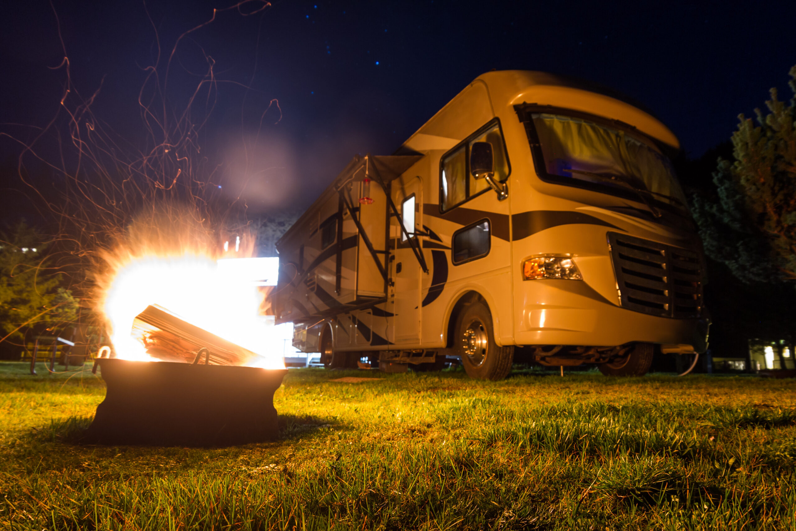 A campfire burns brightly in front of a parked RV at night, with sparks rising into the air. The scene is set on grass, surrounded by trees under a dark, starry sky.
