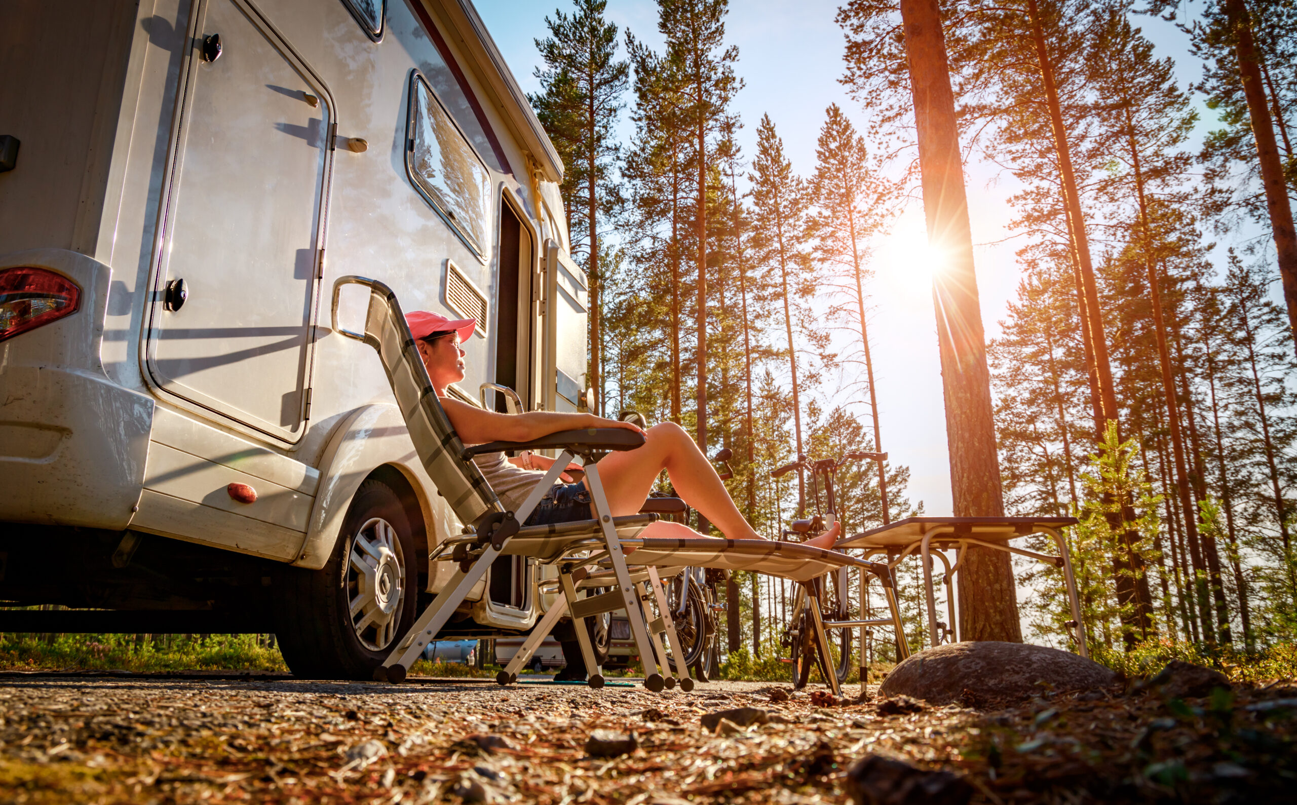 A person relaxes on a lounge chair beside a camper van in a sunlit forest, wearing a hat and sunglasses. Tall pine trees and bright sunlight create a peaceful outdoor atmosphere.