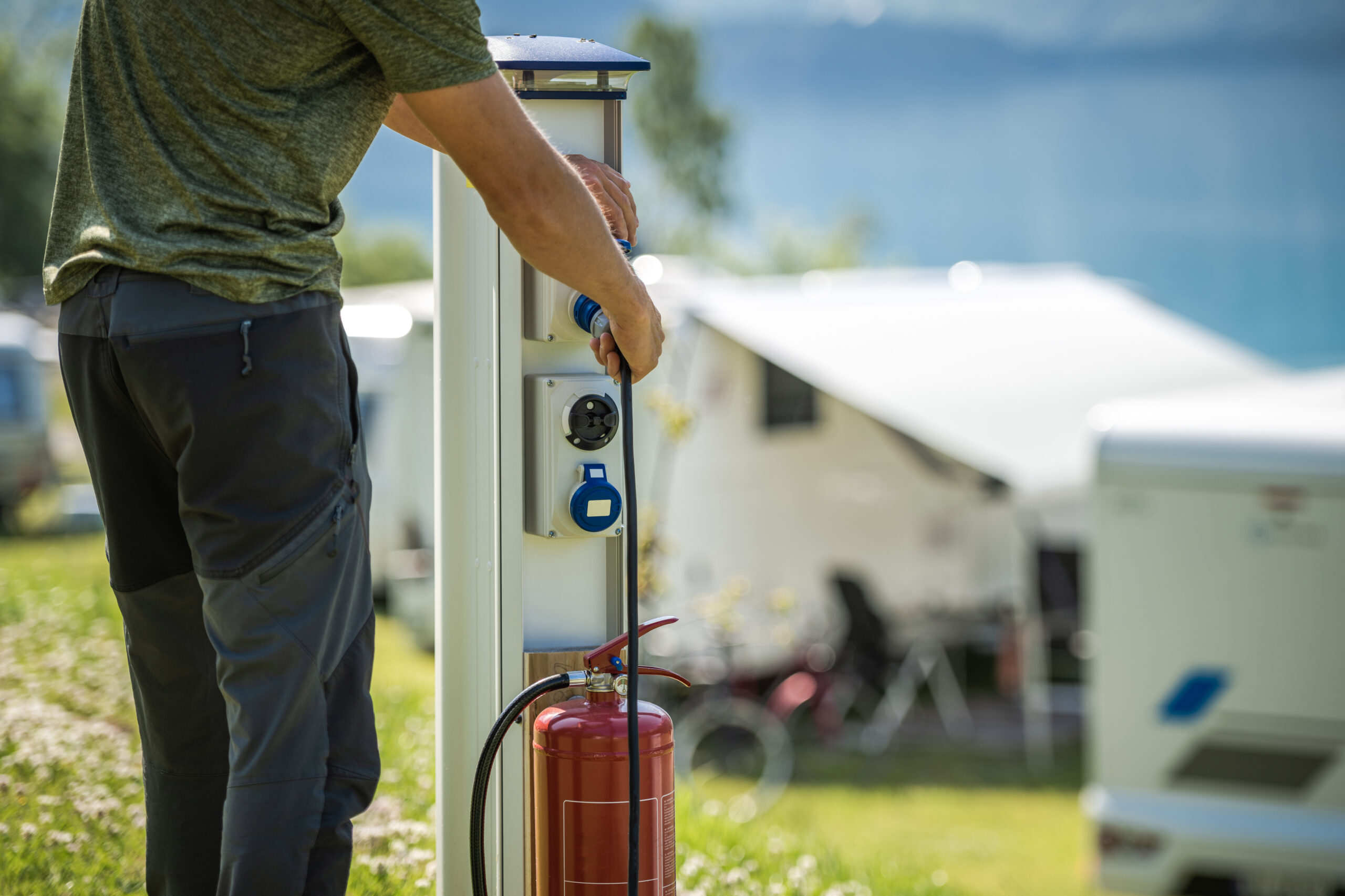 A person plugs an electric cable into a power station at a campsite, with a red fire extinguisher nearby and white camper vans in the blurred background.